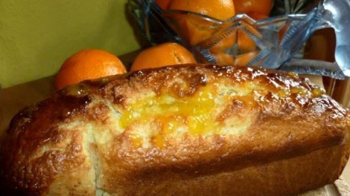 Orangenkuchen gl&auml;nzt frisch auf einem Holzbrett, umgeben von einer Obstschale mit ganzen Orangen im Hintergrund.