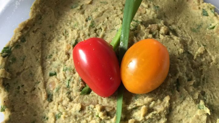 Eine gr&uuml;ne, cremige, vegetarische Brotauflage mit einem Fr&uuml;hlingszwiebel und zwei Kirschtomaten in der Mitte.