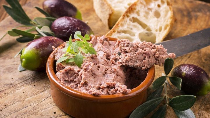 Eine Sch&uuml;ssel mit H&uuml;hnerleberpastete steht auf einem Holzbrett, umgeben von Oliven und Brotst&uuml;cken, bereit zum Servieren.