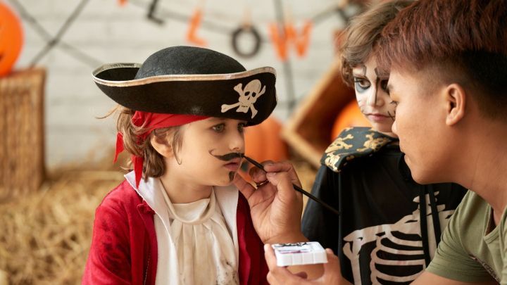 Ein Kind im Piratenkost&uuml;m sitzt ruhig, w&auml;hrend jemand ihm einen Schnurrbart aufmalt, umgeben von festlicher Dekoration.