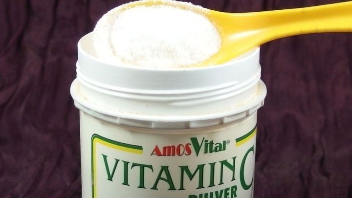 Ein gelber L&ouml;ffel hebt wei&szlig;en Vitamin C-Pulver aus einem Beh&auml;lter, auf einem dunklen Hintergrund.