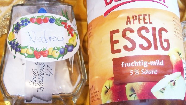 Eine Glasflasche mit der Aufschrift "Natron" steht neben einer Flasche "Apfel Essig", auf einem bunten Untergrund.