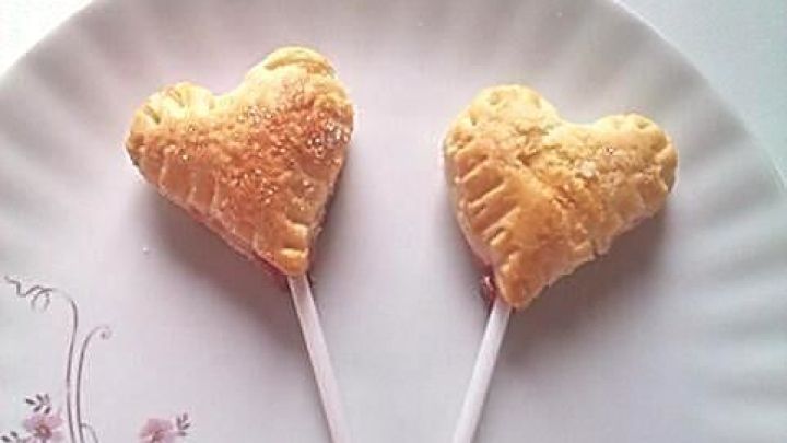 Zwei herzf&ouml;rmige Bl&auml;tterteig-Snacks auf einem wei&szlig;en Teller mit Blumenmuster stehen aufrecht auf Holzst&auml;bchen.