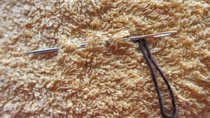Nadel sticht durch braun-gelben Frottee, eine Schlinge zieht sich hindurch. Die Textur zeigt dichte, weiche Fasern.