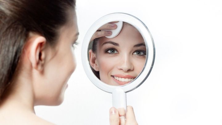 Eine Frau betrachtet sich in einem Handspiegel, w&auml;hrend sie ihr Gesicht sanft reinigt und l&auml;chelt. Fruchts&auml;urepeeling unterst&uuml;tzt die Hautpflege.