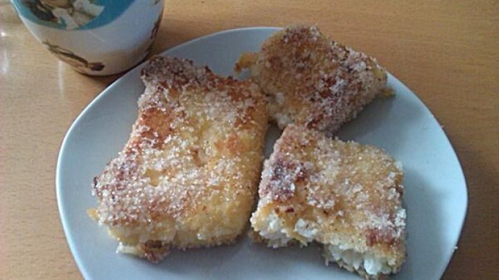 Auf einem wei&szlig;en Teller liegen drei gebratene Milchreisschnitten, daneben steht eine Tasse auf einem Holztisch.