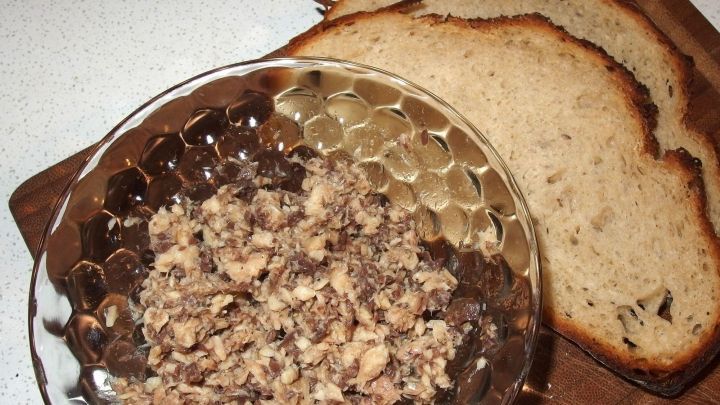 Ein Durchscheinendes Sch&auml;lchen mit zerdr&uuml;ckten &Ouml;lsardinen steht neben mehreren Scheiben Brot auf einem Holzbrett.
