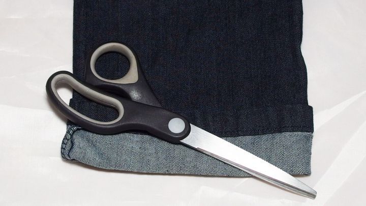 Eine Schere liegt neben dem Originalsaum einer dunkelblauen Jeans, bereit, um diese zu k&uuml;rzen.