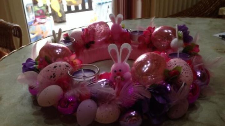 Ein bunter Osterkranz mit Eiern und Kerzen auf einem Tisch; umgeben von Fr&uuml;hlingsdeko in einem gem&uuml;tlichen Raum.
