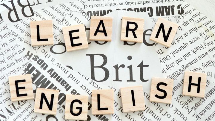 Holzbuchstaben formen den Text "LEARN ENGLISH" auf einem bunten Hintergrund aus Zeitungsblasen und Grafiken, ideal zum Vokabeln lernen.