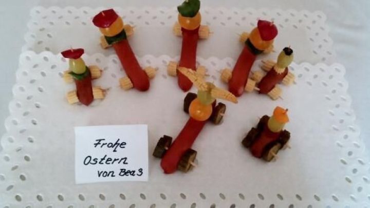 Bunte W&uuml;rstchen-Rennwagen stehen auf einem Tablett, umgeben von festlicher Dekoration f&uuml;r einen Kindergeburtstag. Fr&ouml;hliche Osterw&uuml;nsche sind auf einer Karte platziert.