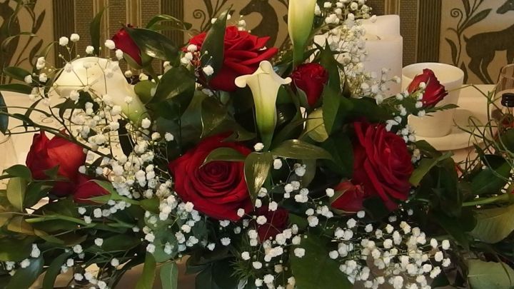 Ein &uuml;ppiger Brautstrau&szlig; aus roten Rosen, wei&szlig;en Lilien und zartem Baby-Atmen auf einem festlich gedeckten Tisch.