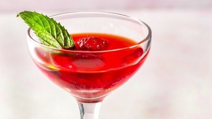 Ein elegantes Glas mit Himbeerlik&ouml;r, garniert mit einem Minzblatt, steht auf einer helle Oberfl&auml;che neben Eisw&uuml;rfeln.