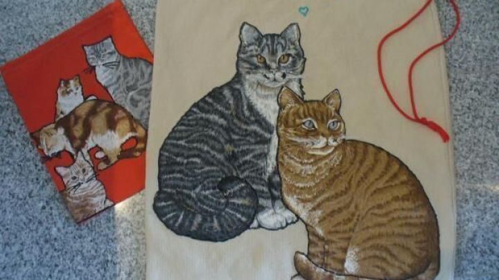 Eine Stofftasche zeigt zwei sitzende Katzen, eine grau und eine orange, in einer gem&uuml;tlichen Umgebung, umgeben von einer weiteren roten Tasche.