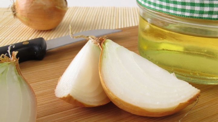 Auf einem Holzbrett liegen eine halbierte Zwiebel und eine ganze Zwiebel, neben einem Glas Zwiebelsaft.