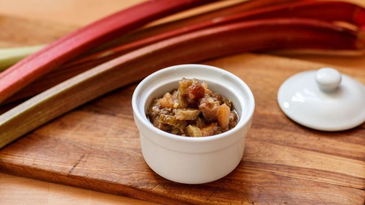Ein Sch&auml;lchen mit Rhabarber-Chutney steht auf einem Holzbrett, umgeben von frischen Rhabarberstangen und einem kleinen Deckel.
