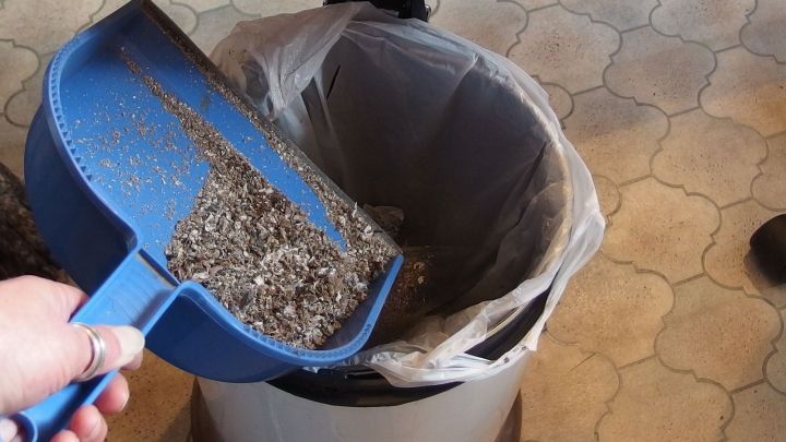 Eine Hand h&auml;lt eine blaue Schaufel, die Abf&auml;lle in einen M&uuml;lleimer leert, der in einer K&uuml;che steht.