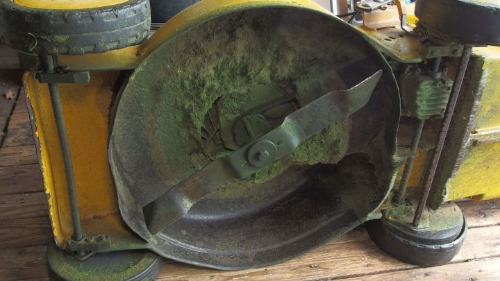 Ein Rasenm&auml;her liegt auf der Seite, w&auml;hrend Grasreste entfernt werden, in einer Werkstatt mit Holzfu&szlig;boden.