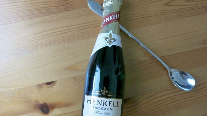 Ein Silberl&ouml;ffel steckt in einer ge&ouml;ffneten Henkell Sektflasche, die auf einem Holztisch liegt.