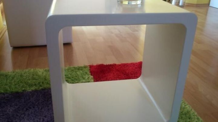 Ein wei&szlig;er Lacktisch steht auf buntem Teppichboden, oben drauf ein Glas mit Kerze, im Hintergrund helle Holzb&ouml;den.