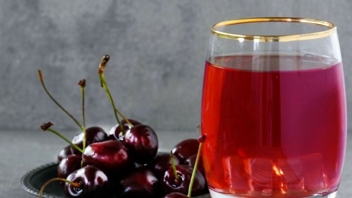 Ein Glas Sauerkirschsaft steht auf einem Tisch, umgeben von frischen Kirschen auf einem Teller in grauer Umgebung.