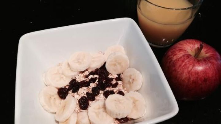 M&uuml;sli mit Bananenscheiben und Rosinen in wei&szlig;er Schale auf schwarzem Tisch, daneben Apfel und Glas Orangensaft.
