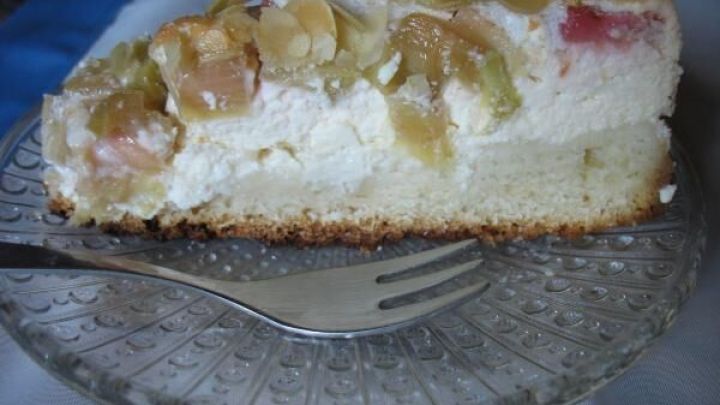 Eine fruchtige Quark-Rhabarber-Torte mit Mandelbaiser liegt auf einem transparenten Teller und wird mit einer Gabel serviert.