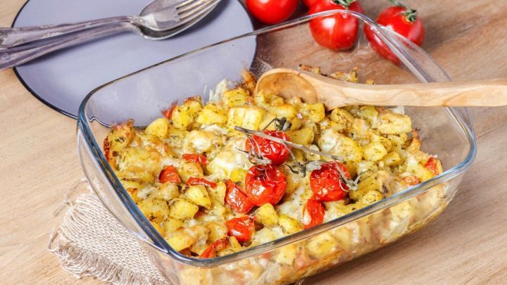 Ein bunter Kartoffel-Tomaten-K&auml;se-Auflauf liegt in einer Glasform auf einem Holztisch, umgeben von frischen Tomaten und dekorativen Vasen.