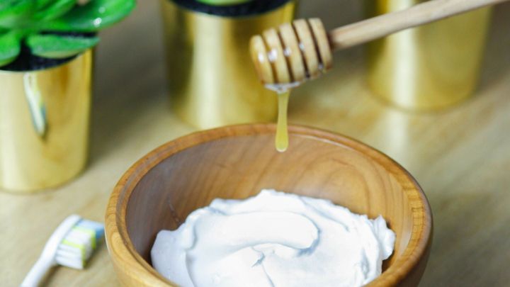 Eine Holzsch&uuml;ssel mit Cremes wird mit Honig bef&uuml;llt, w&auml;hrend ein Zahnb&uuml;rste und Pflanzen im Hintergrund stehen.