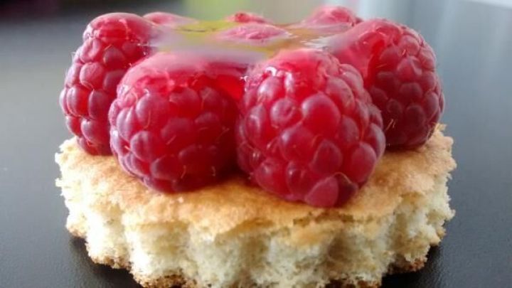 T&ouml;rtchen mit saftigen Himbeeren belegt auf einem knusprigen Biskuitboden, pr&auml;sentiert auf einem dunklen, glatten Tisch.