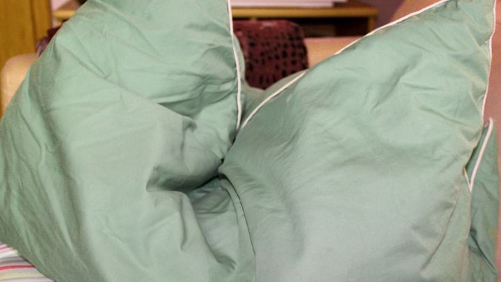 Zwei gr&uuml;ne Federkissen liegen auf einem Bett, w&auml;hrend im Hintergrund eine gem&uuml;tliche Wohnzimmereinrichtung sichtbar ist.