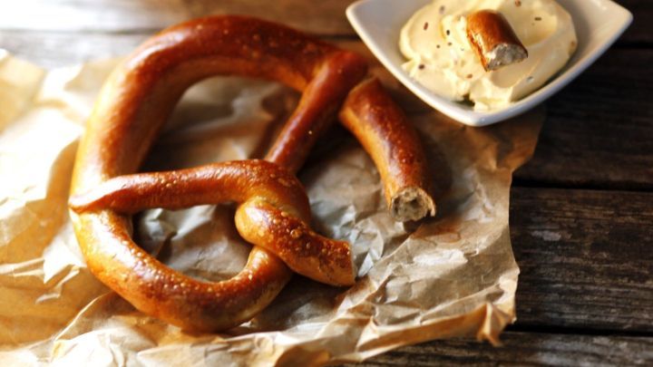 Ein frisch gebackenes Brezel liegt auf braunem Papier, neben einem Sch&auml;lchen mit cremigem Obazda auf einem Holztisch.
