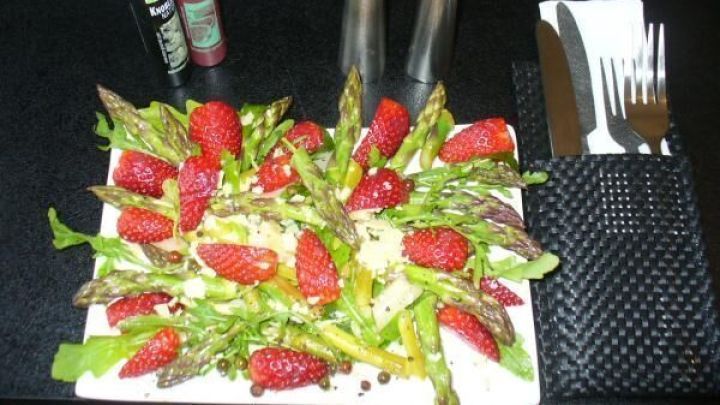 Spargelsalat mit Erdbeeren liegt auf einem Teller, umgeben von Besteck, Pfeffer- und Salzstreuer und Dressing-Flaschen.