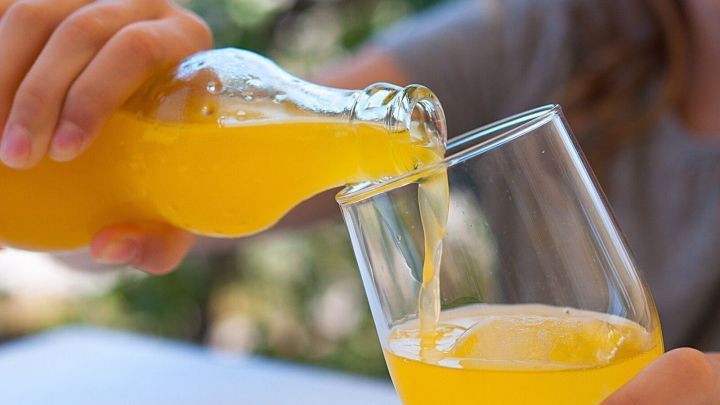 Eine Hand gie&szlig;t ein orangefarbenes Getr&auml;nk aus einer Flasche in ein Glas, im Hintergrund unscharfe gr&uuml;ne Natur.