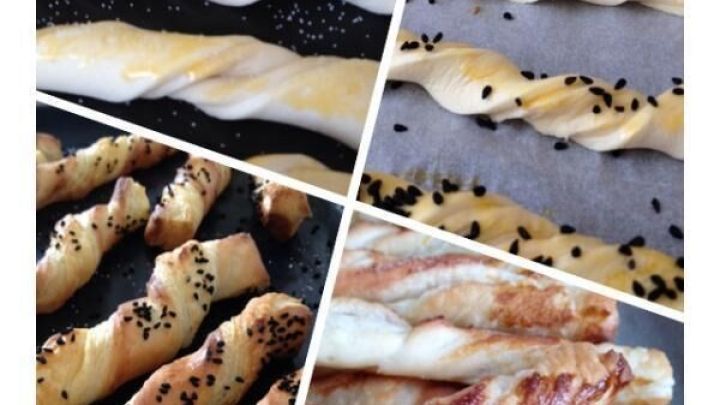 Bl&auml;tterteig-Stangen liegen gedreht auf Backblechen, teils ungebacken, teils golden mit schwarzem Sesam bestreut.