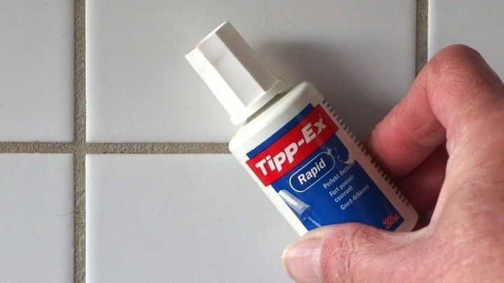 Eine Hand h&auml;lt eine Tipp-Ex-Flasche, die auf eine schmutzige Fuge zwischen wei&szlig;en Fliesen gerichtet ist.