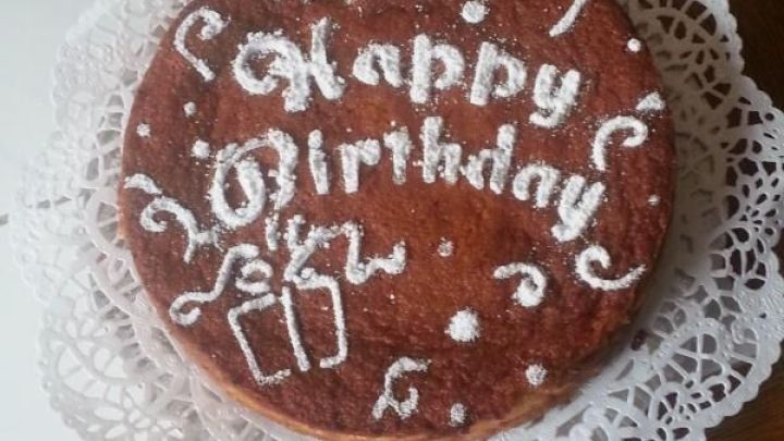 K&auml;sekuchen liegt auf einem filigranen Untersetzer und zeigt "Happy Birthday" mit Dekoration in einer festlichen Umgebung.
