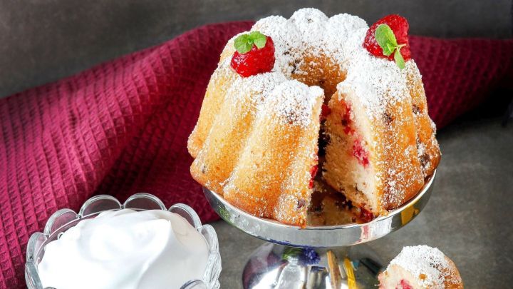 Ein Mini-Himbeer-Gugl steht auf einem silbernen Podest, umgeben von Sahne und fliederfarbenen Blumen, leicht gepudert mit Zucker.