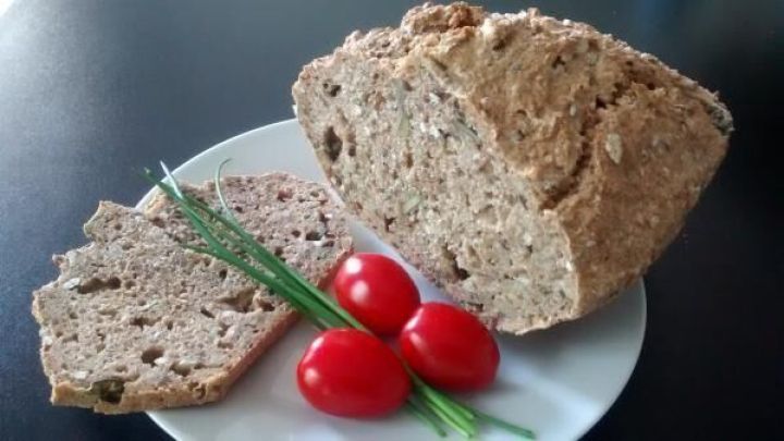 Ein Laib Brot liegt auf einem Teller mit Tomaten und Schnittlauch, daneben zwei geschnittene Scheiben auf einem dunklen Tisch.