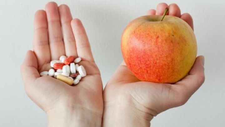 Eine Hand zeigt eine Auswahl an Nahrungserg&auml;nzungsmitteln, w&auml;hrend die andere Hand einen frischen Apfel h&auml;lt, was einen Vergleich von Vitaminpr&auml;paraten und nat&uuml;rlichen Lebensmitteln darstellt.