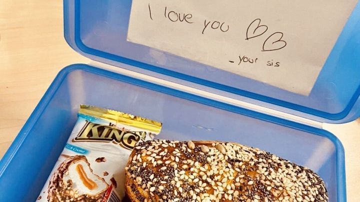 In einer blauen Brotdose steht ein Br&ouml;tchen, neben dem ein Zettel mit liebevollen Botschaften und Snacks liegt.