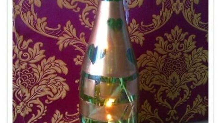 Weinflasche mit Lichterkette als Deko, umwickelt mit gr&uuml;nem Band, steht vor r&ouml;tlich-goldenem, ornamentalen Hintergrund.