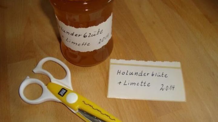 Ein Marmeladenglas mit gelbem Deckel steht auf einem Tisch, beschriftet mit "Holunderbl&uuml;te + Limette 2014". Eine gelbe Zickzackschere und ein Etikett mit derselben Aufschrift liegen daneben.