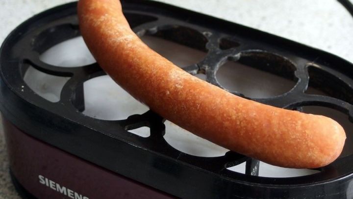 Ein W&uuml;rstchen liegt auf einem Eierkocher, w&auml;hrend es erhitzt wird. Der K&uuml;chenuntergrund ist hell und modern.