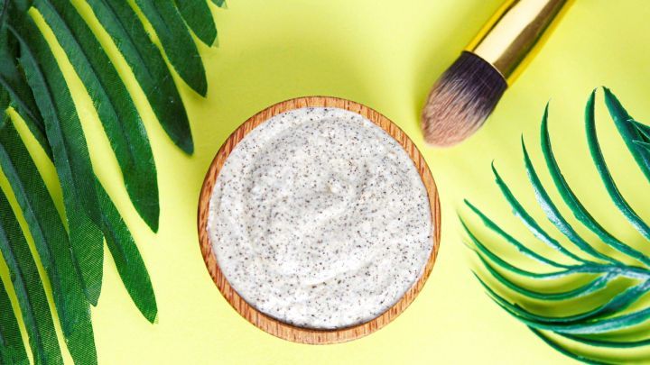 Eine Gesichtsmaske in einer Holzsch&uuml;ssel wird neben einem Pinsel und gr&uuml;nen Bl&auml;ttern auf einem gelben Untergrund pr&auml;sentiert.