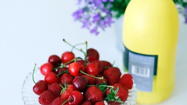 Eine Sch&uuml;ssel mit frischen Beeren steht auf einem Tisch, w&auml;hrend daneben Flasche mit Zitronensaft und Blumen steht.