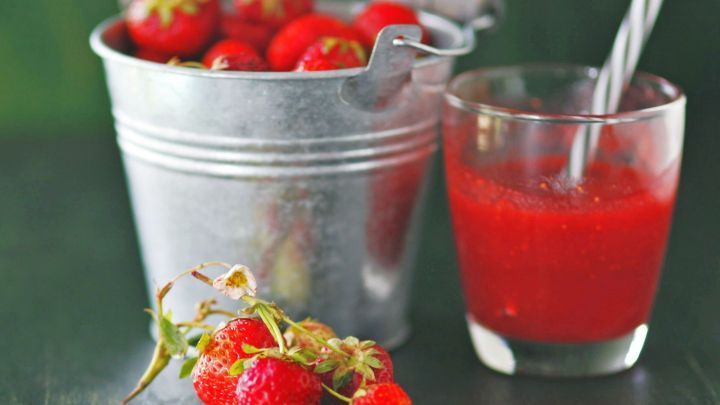 Ein Eimer mit frischen Erdbeeren steht neben einem Glas mit Erdbeerlik&ouml;r, alles vor einem gr&uuml;nen Hintergrund.