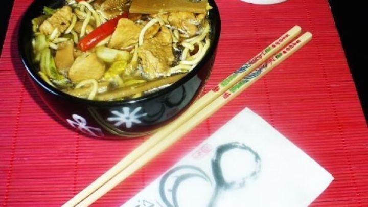 In einer Sch&uuml;ssel mit scharfer chinesischer H&uuml;hnernudelsuppe liegen frische Zutaten, w&auml;hrend Gl&uuml;cks-Katze im Hintergrund steht.