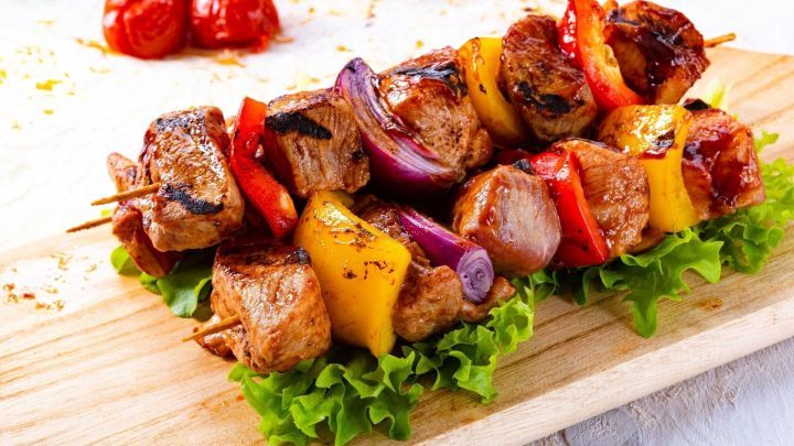 Saftige Schaschlikst&uuml;cke liegen auf einem Holzbrett, umgeben von buntem Gem&uuml;se und frischem Salat.