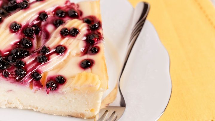 Eine St&uuml;ck Heidelbeer-Schmand-Torte liegt auf einem wei&szlig;en Teller, garniert mit schwarzen Beeren und einer Gabel daneben.